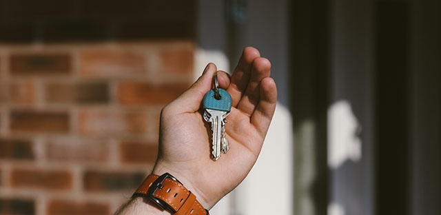 Image of Hand holding keys in front of a house after acquiring property